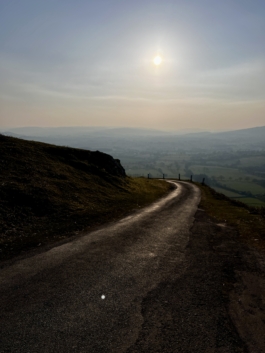 Single width country road, winding around the corner, with fields in the background with the sun in the sky. Ross Woodhall - Action & Lifestyle Photography